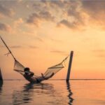 man hanging in hammock over water between posts and without trees